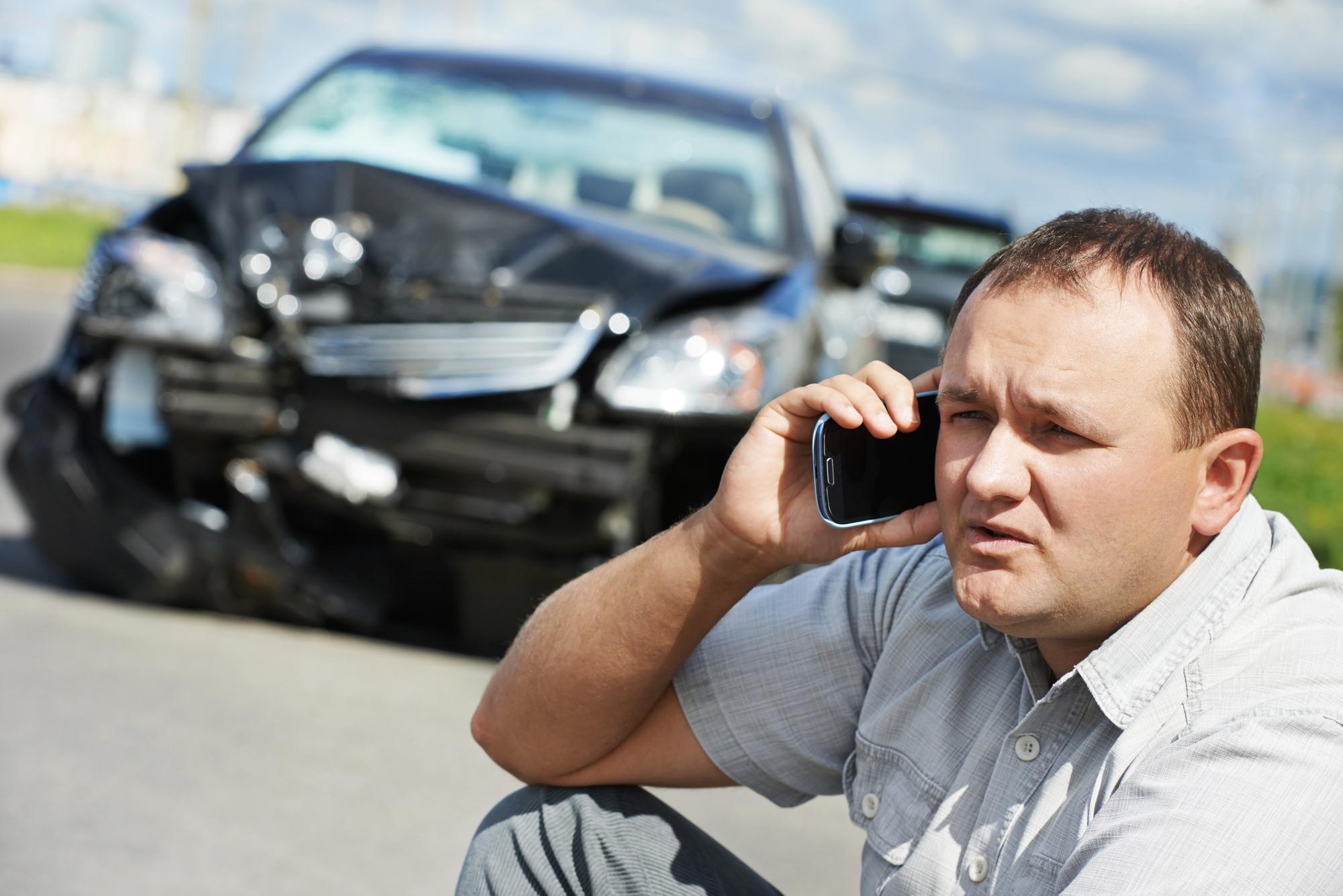 A man sitting beside a wrecked car, preparing to describe a car accident to an insurance agent on the phone.