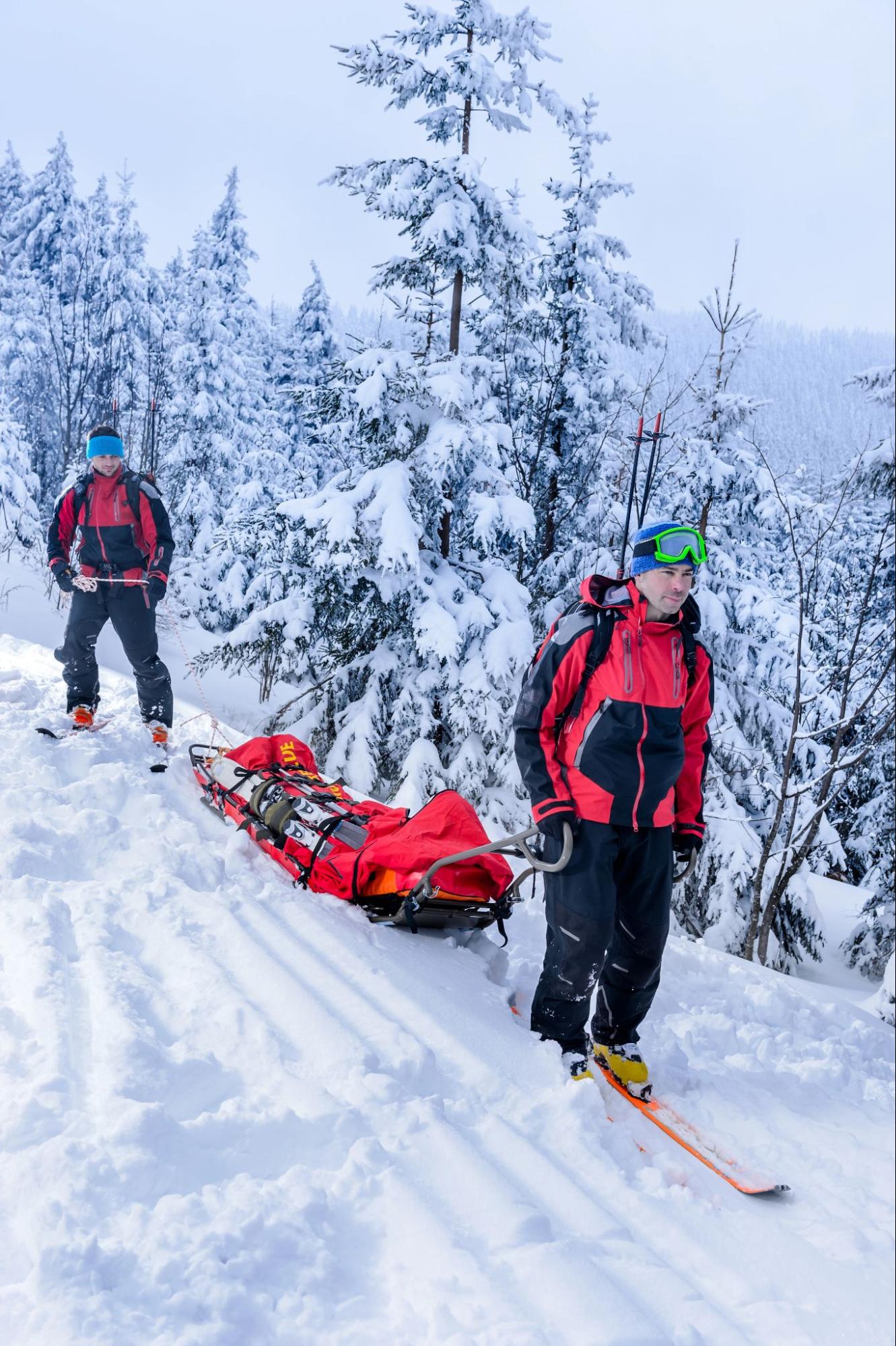 Ski patrol rescuers transport a man with ski injuries on a rescue sled through a snow-covered mountain forest.