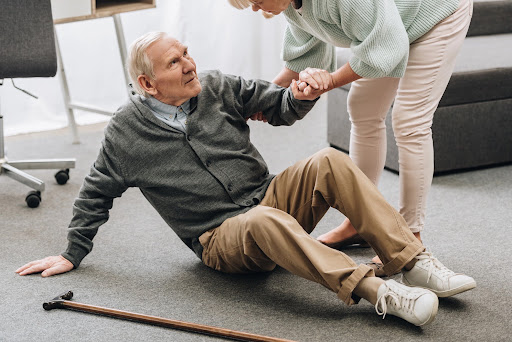 An elderly man with a cane sits on the floor after a slip and fall accident while a woman helps him stand up.